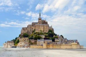 Vacances familiales pas chères au Mont Saint Michel. On aperçoit le mont sur la plage, le ciel est bleu et nuageux.