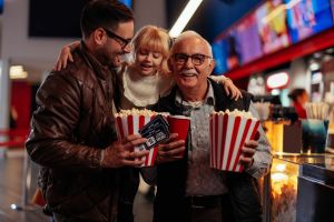 Cinéma pas cher : un grand-père, son fils et sa petite-fille vont au cinéma en souriant car ils profitent du moment avec du pop-corn.