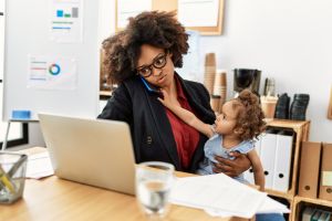 Une jeune femme de couleur en train de travailler par téléphone devant son ordinateur avec son enfant dans les bras.