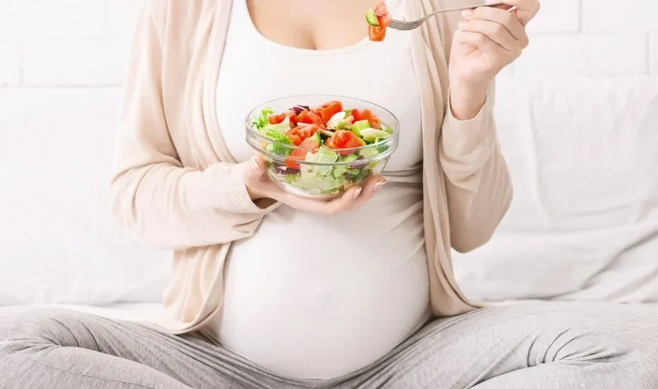 Une femme enceinte mange une salade assise sur son lit.