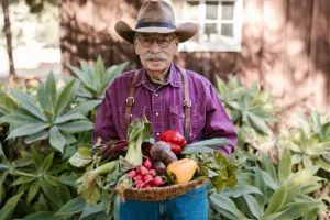 Un agriculteur qui tient des légumes dans ses mains. Il porte un chapeau et une chemise rose.