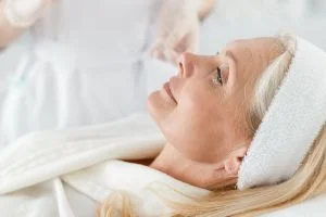 Une femme senior est couchée dans un institut en peignoir. Sa peau est resplendissante, elle sourit.