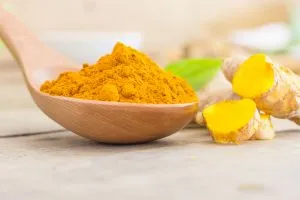 Une cuillère en bois remplie de poudre de curcuma riche en curcumine posée sur une table à côté de racine de curcuma.