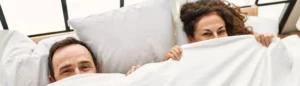 Un couple dans lit qui se cache sous une couette après avoir réglé le problème d'érection grâce à une solution naturelle trouvée en pharmacie.