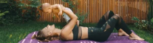 Une jeune maman allongée dans son jardin en tenu de sport qui fait du yoga postnatal avec son bébé dans les bras.