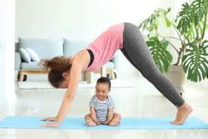 Une jeune maman qui fait une posture de yoga post-natal avec son bébé assis sur le tapis.