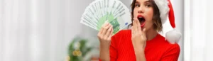 Une jeune femme porte un bonnet de Noël et tient des billets en main qui correspondent à la prime de Noël 2025.