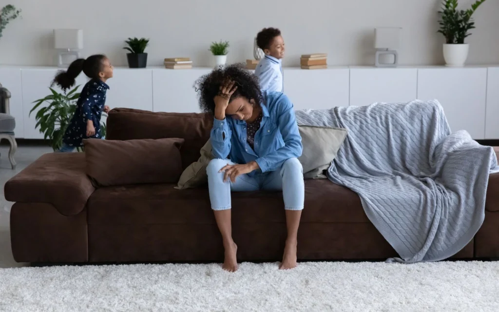 Une maman fatiguée assise sur son canapé, ses enfants courent dans le salon.