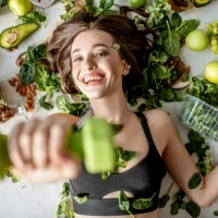 Une jeune femme qui soulève des haltères entourée de fruits et légumes bons pour la nutrition sportive.